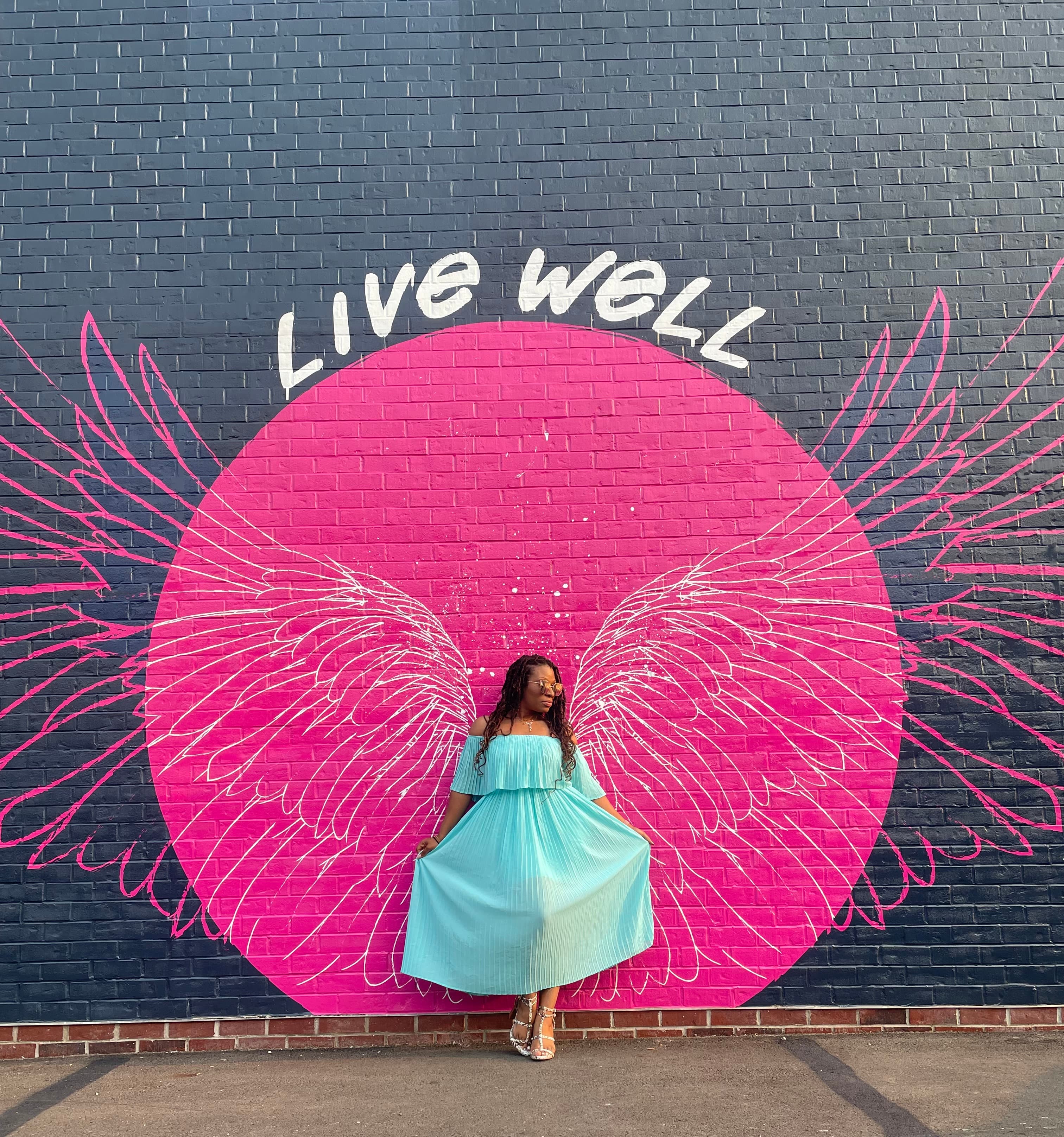 Travel advisor Libby posing in a blue dress against a wall with bright pink circle and wings and 'Live Well' sign