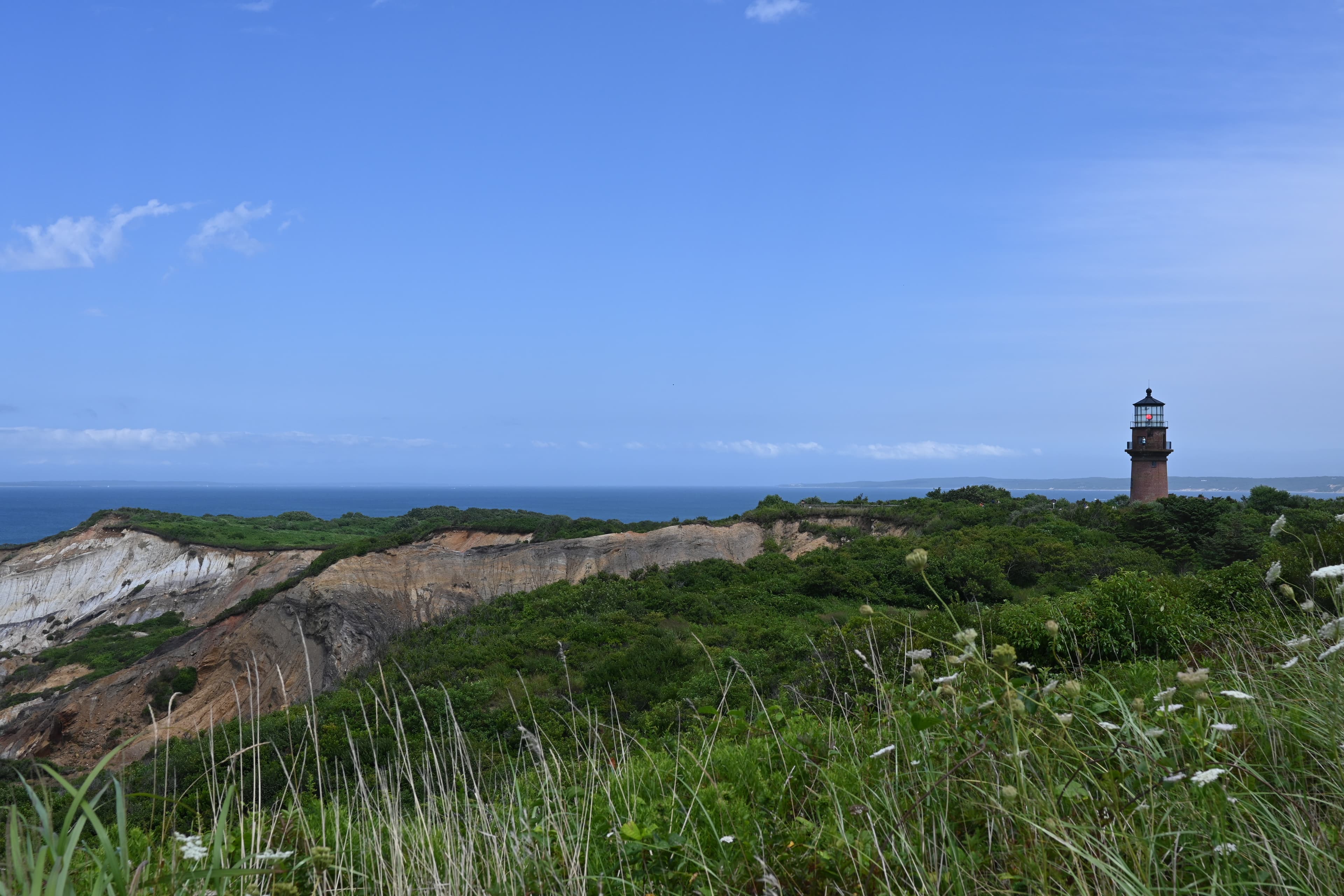 A view of a green meadow with cliffs on a sunny day overloooking the ocean with a lighthouse in view.