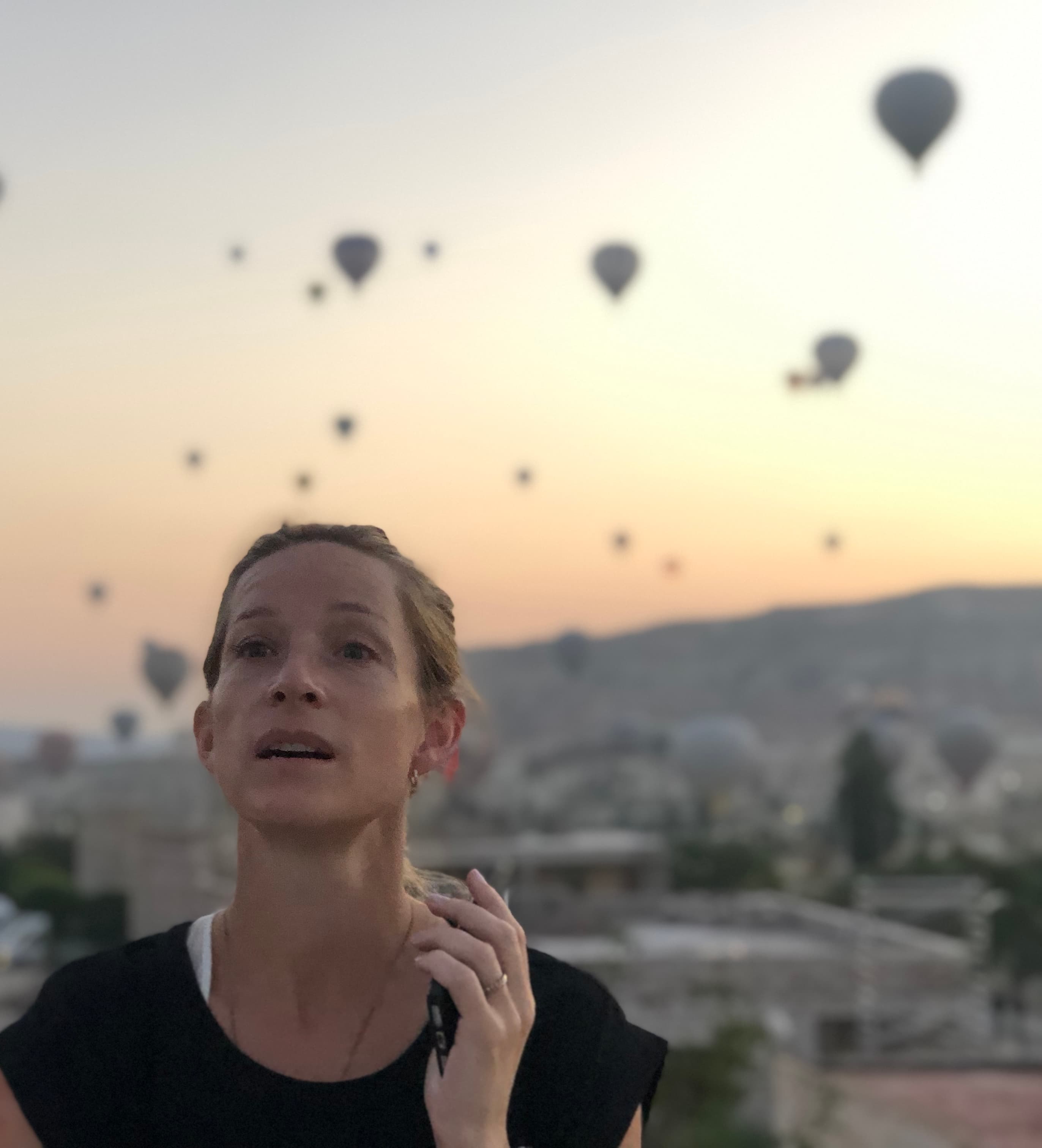 A person standing in front of a view of hot air balloons in the sky with a sunrise in the background.