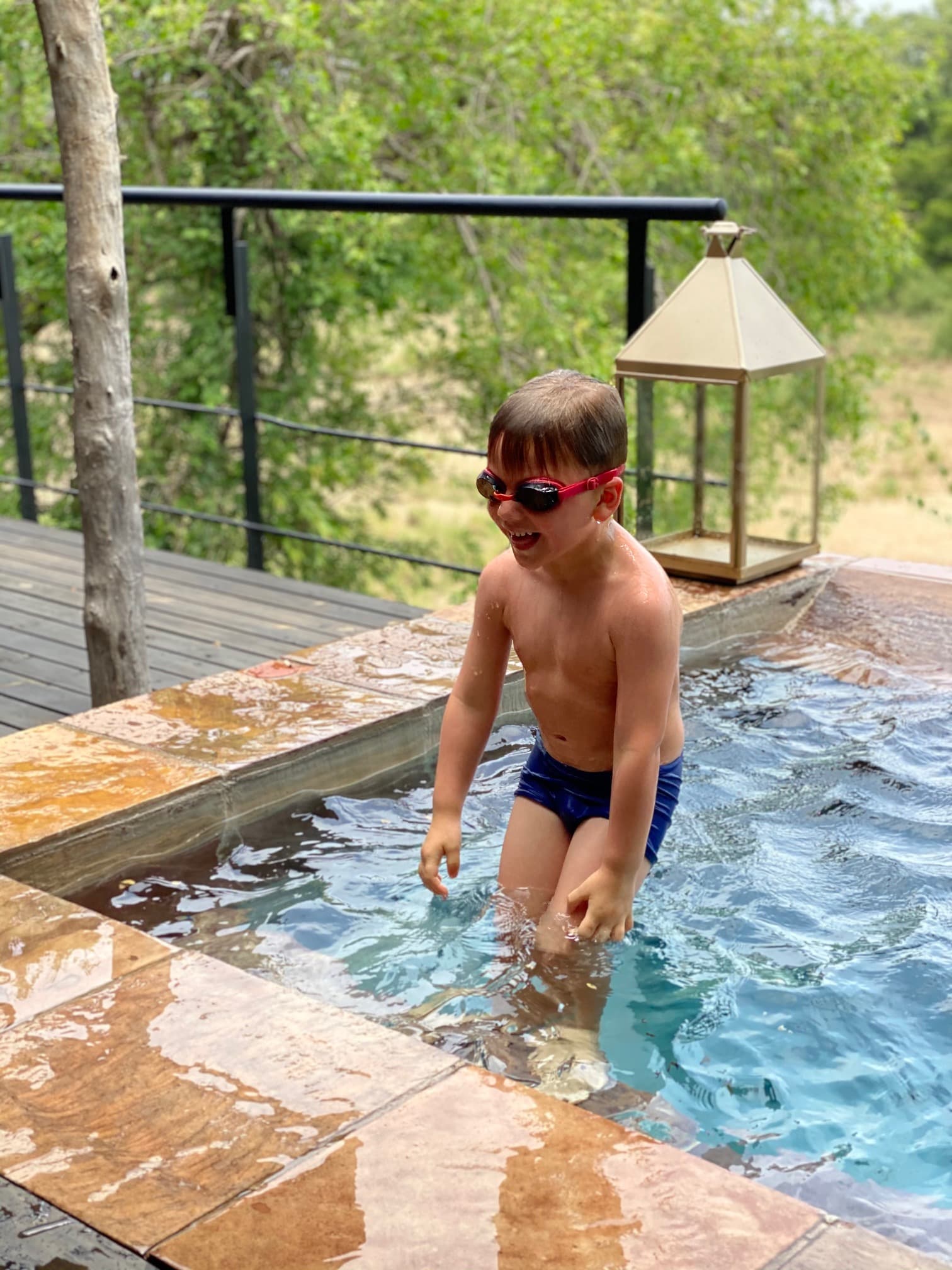 A kid wearing goggles and swim trunks jumping into an outdoor pool. There is a wooden deck, black railing and metal lantern in the surrounding area, as well as trees in the background.