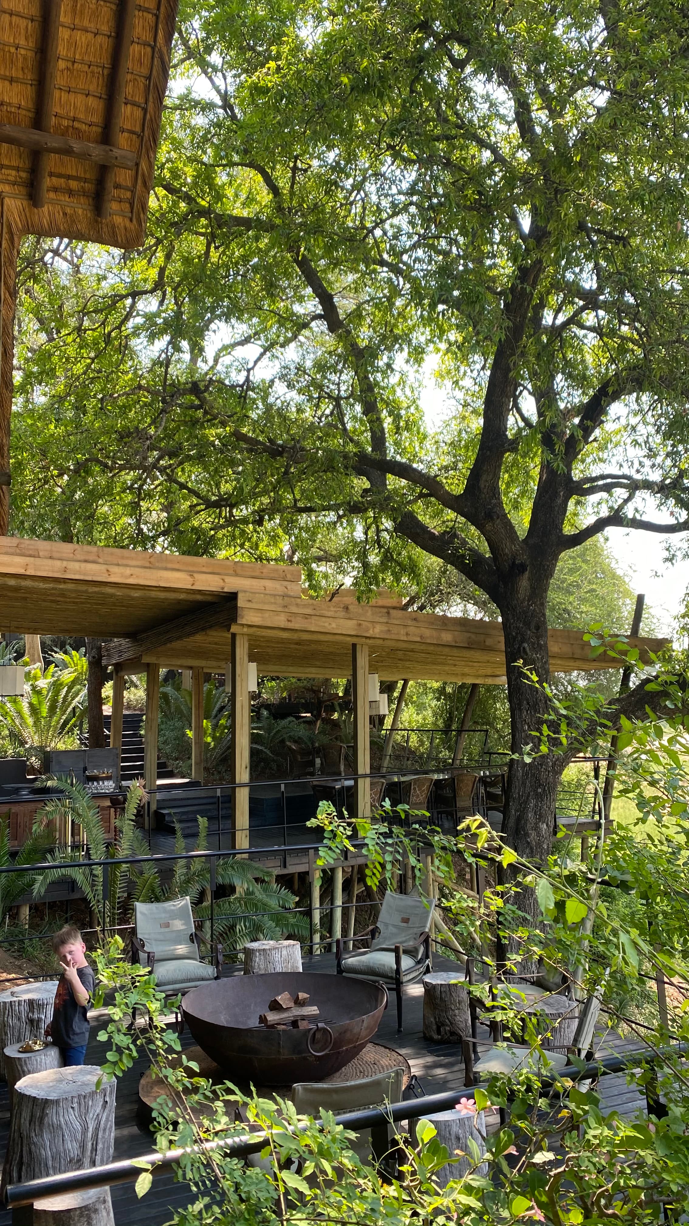 A beautiful view of an outdoor patio surrounded by lush green trees, lounge chairs, a stone fireplace and a wooden roof covering.