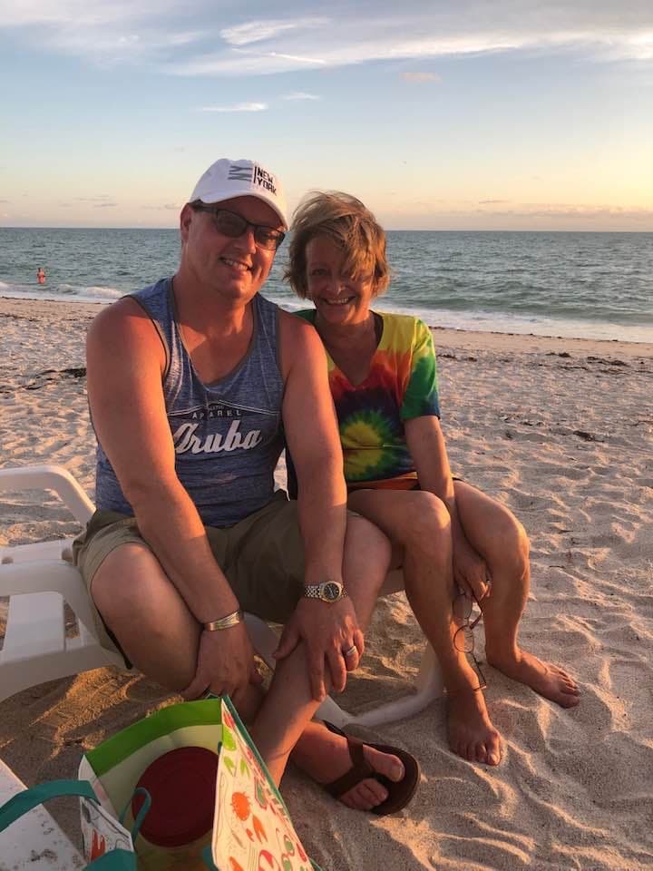 Barry in a tank top and shorts sitting next to a woman in a tie-dye shirt on a lounge chair on a beach at sunset.
