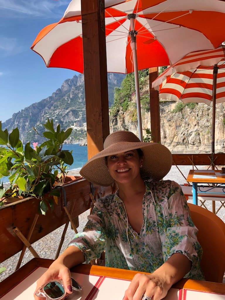 Travel advisor Catherine sitting at a table at Arienzo beach club in a hat and green and white shirt. There are red and white umbrellas behind and the ocean in view.