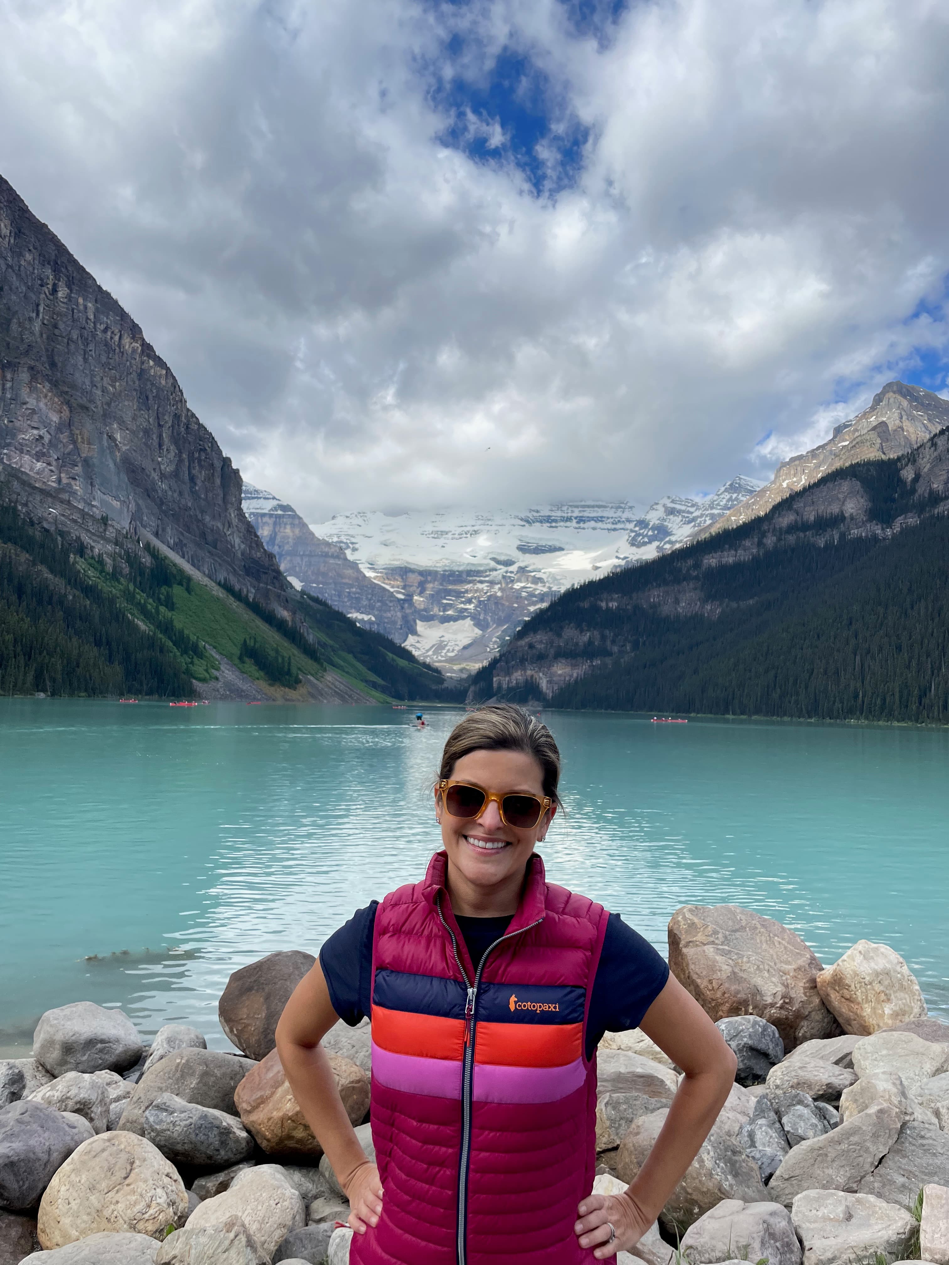 Travel advisor Catherine in a colorful pink vest standing in front of a beautiful lake with mountains in view