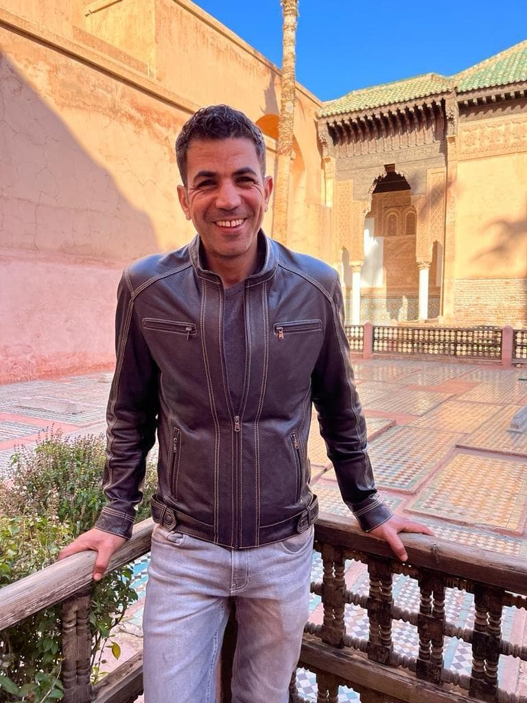 Hassane smiling in a brown leather jacket and standing on a balcony in a Middle Eastern-style plaza.