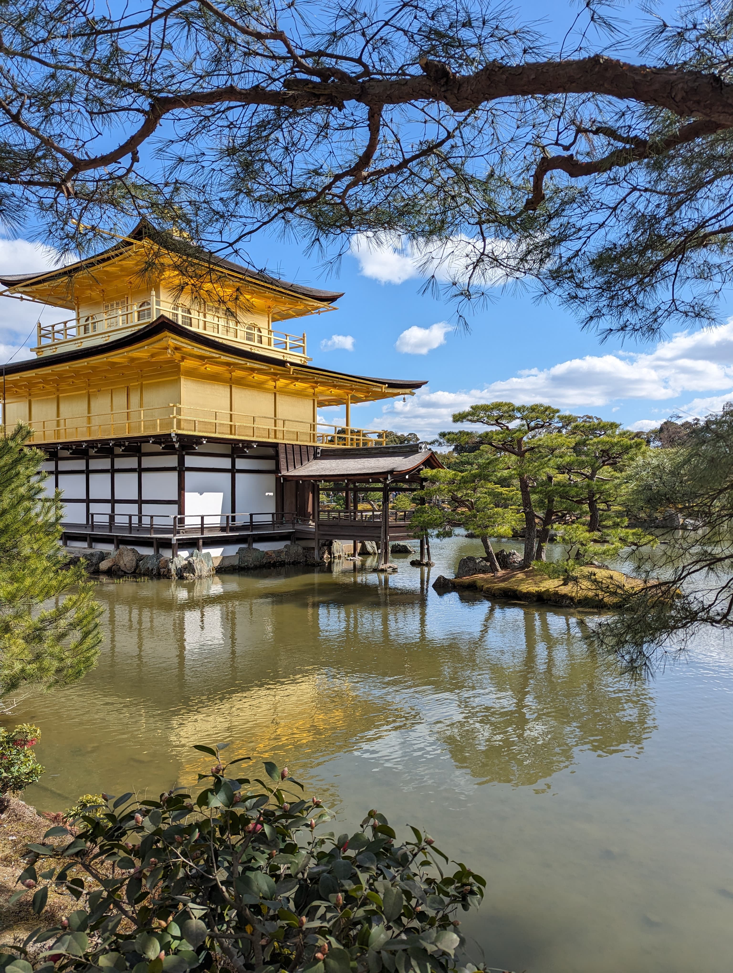 A beautiful yellow temple near a lake outside. There are trees and rocks in the surrounding area.