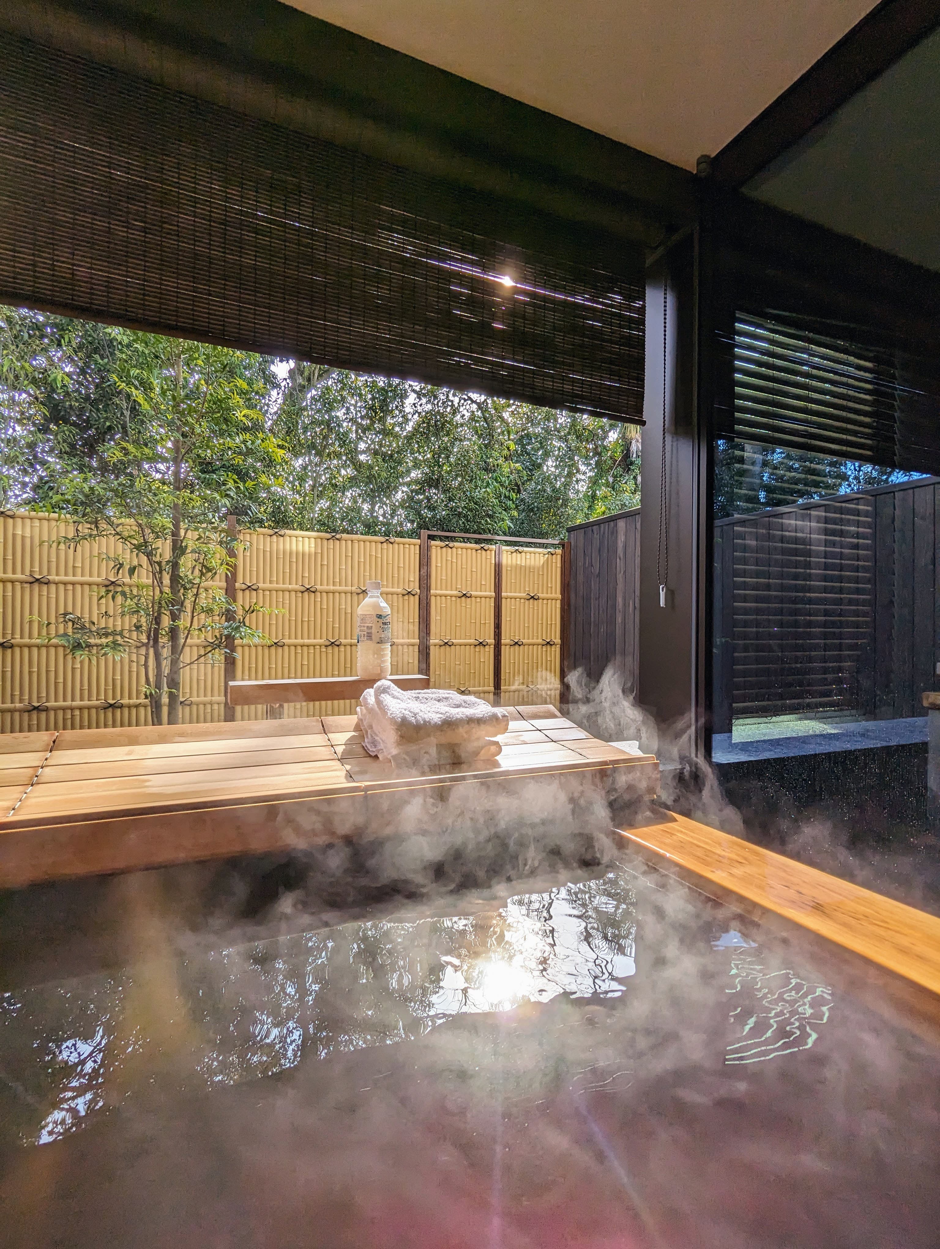 A view of a steam bath surrounded wooden architecture, towels and a fence surrounded by trees.