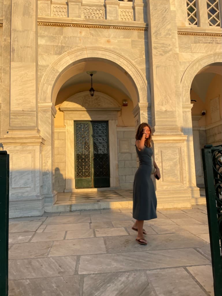 A woman wearing a black dress and posing in front of a beautiful old stone building with archways illuminated at sunset.