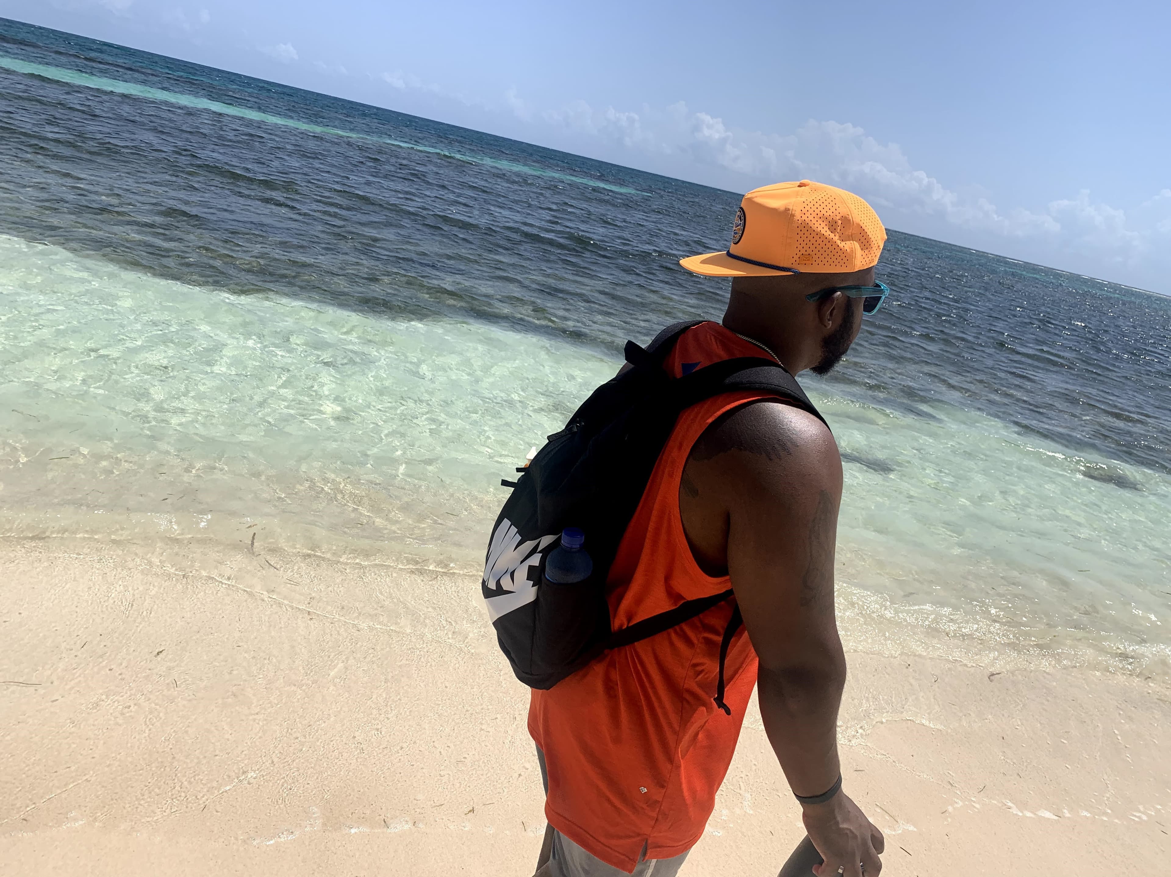 Picture of Harold wearing a tank top and orange hat walking on a beach with crystal clear water