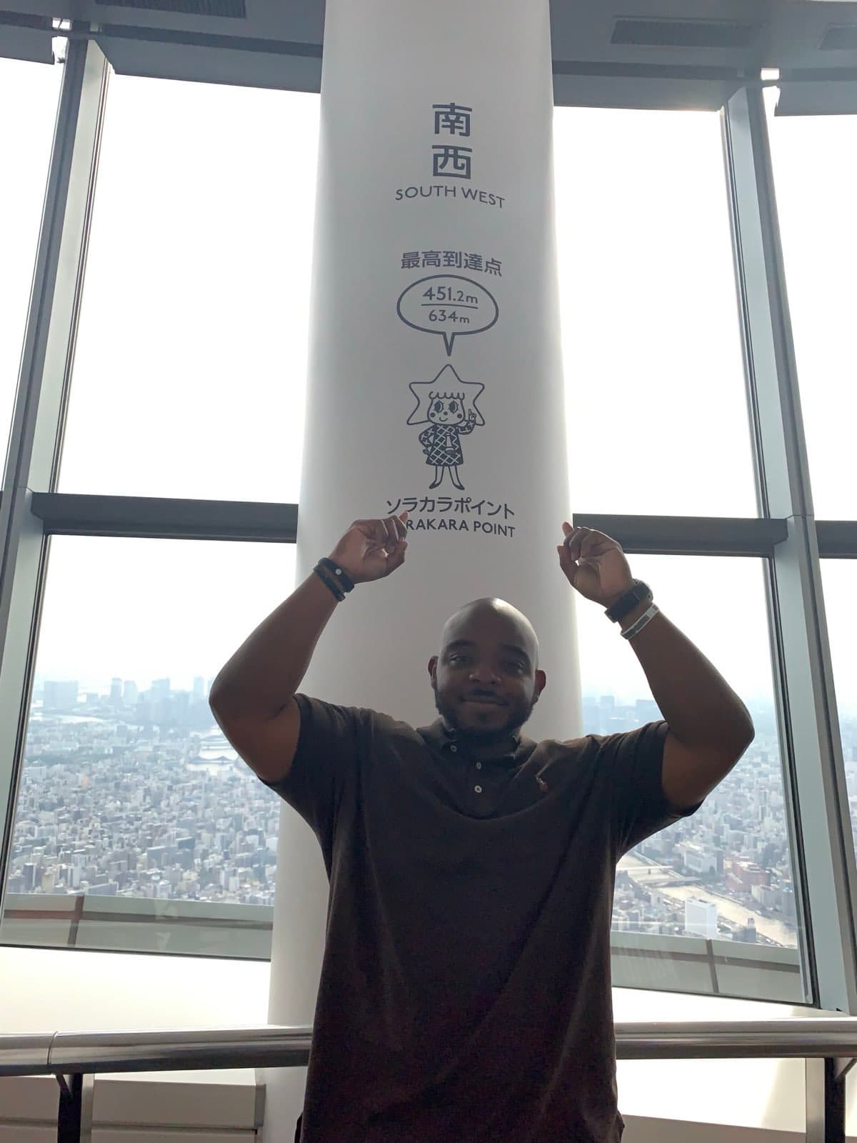 Picture of Harold standing in front of a pole with Japanese writing on it with the view of a skyline in the background.