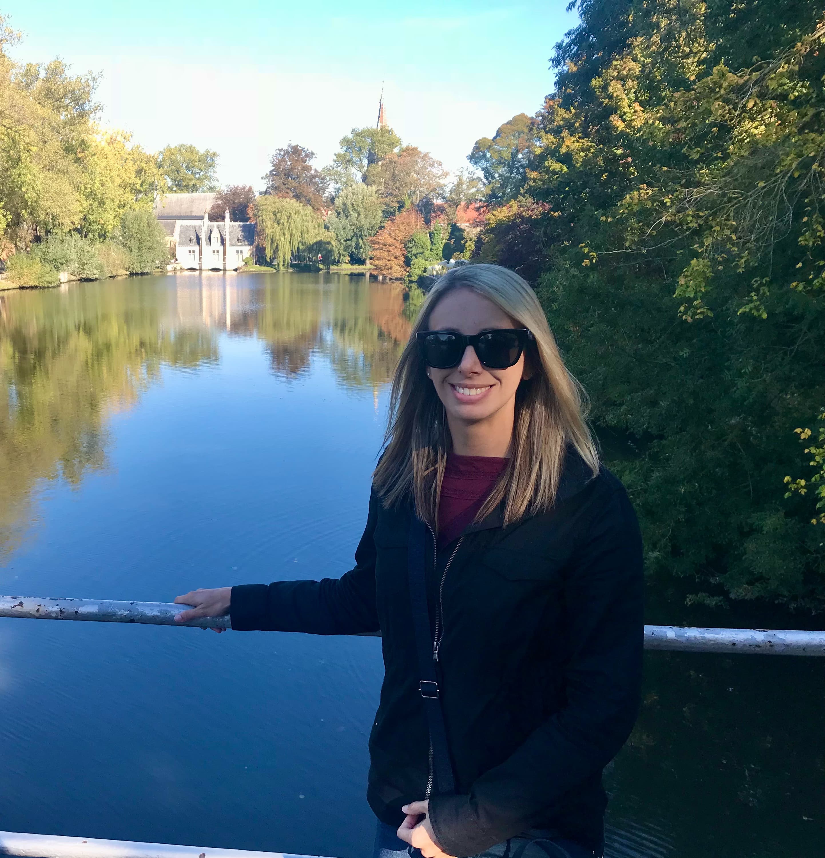 Leslie posing at a railing on a walkway above a waterway with foliage and historical buildings in the distance.