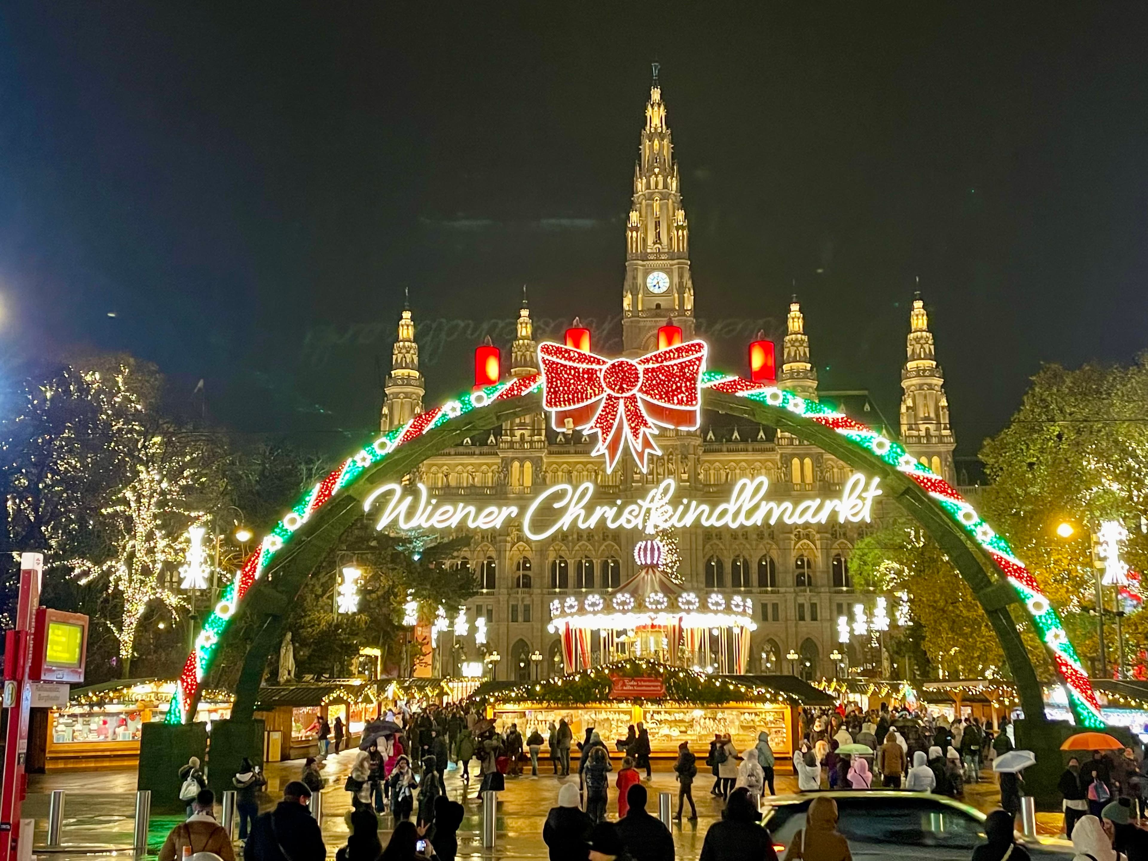 An outdoor view of a European Christmas market with various lights at night and people walking around in the surrounding areas.
