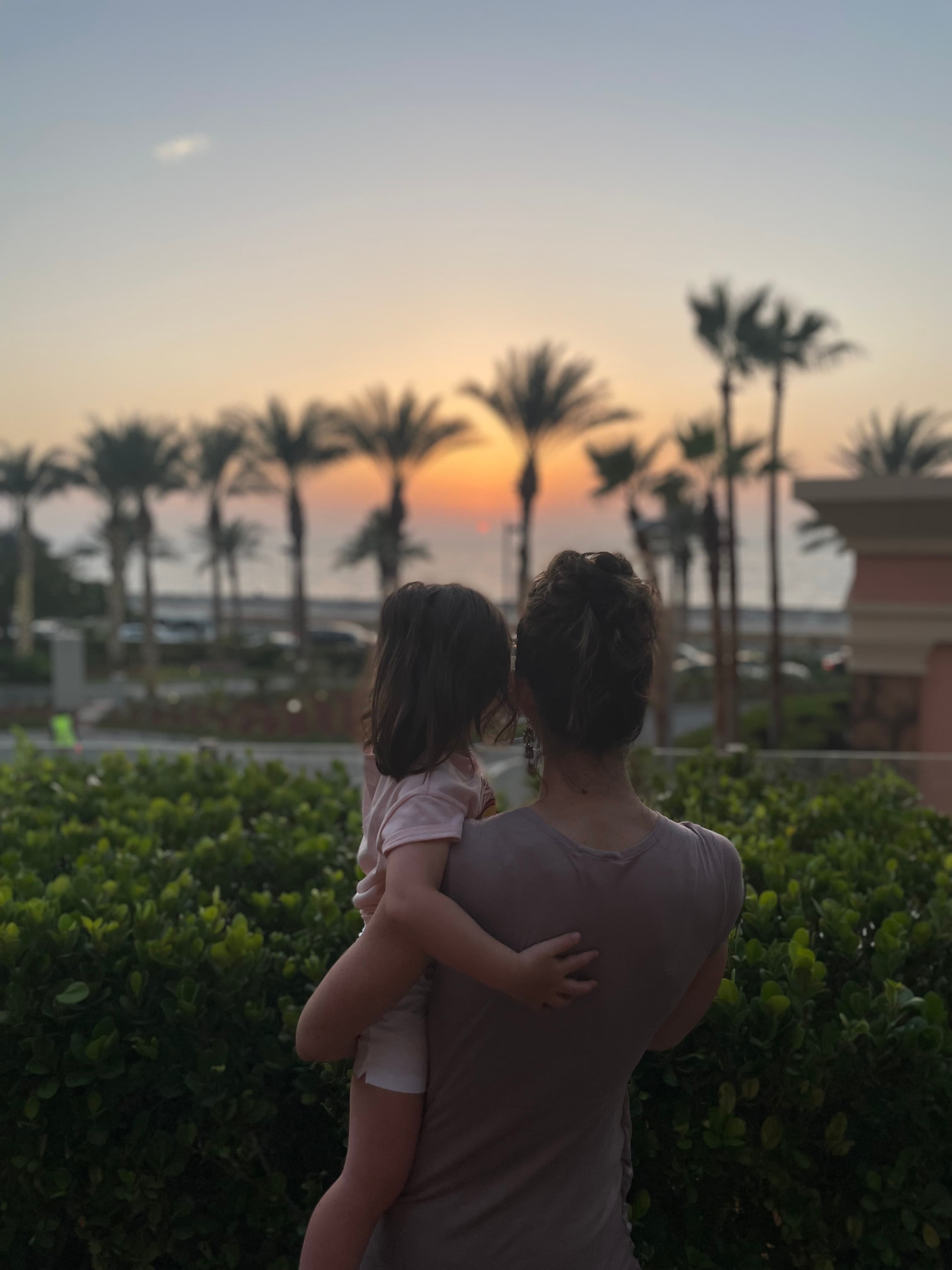 A photo of Jennifer holding her daughter and looking out toward palm trees, bushes and an orange sunset.