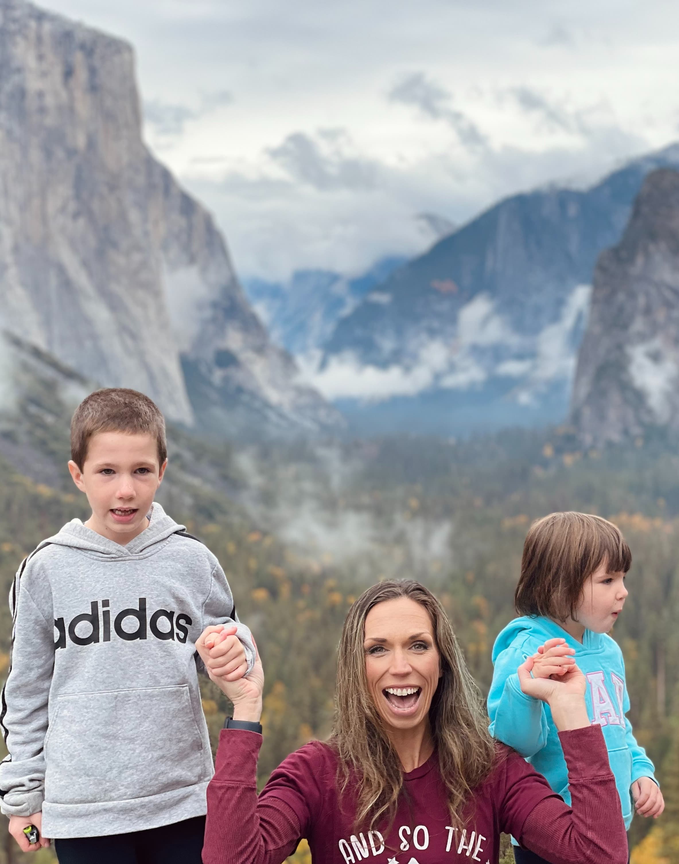 A photo of Jennifer smiling and holding her childrens hands in front of a forest and large mountains.