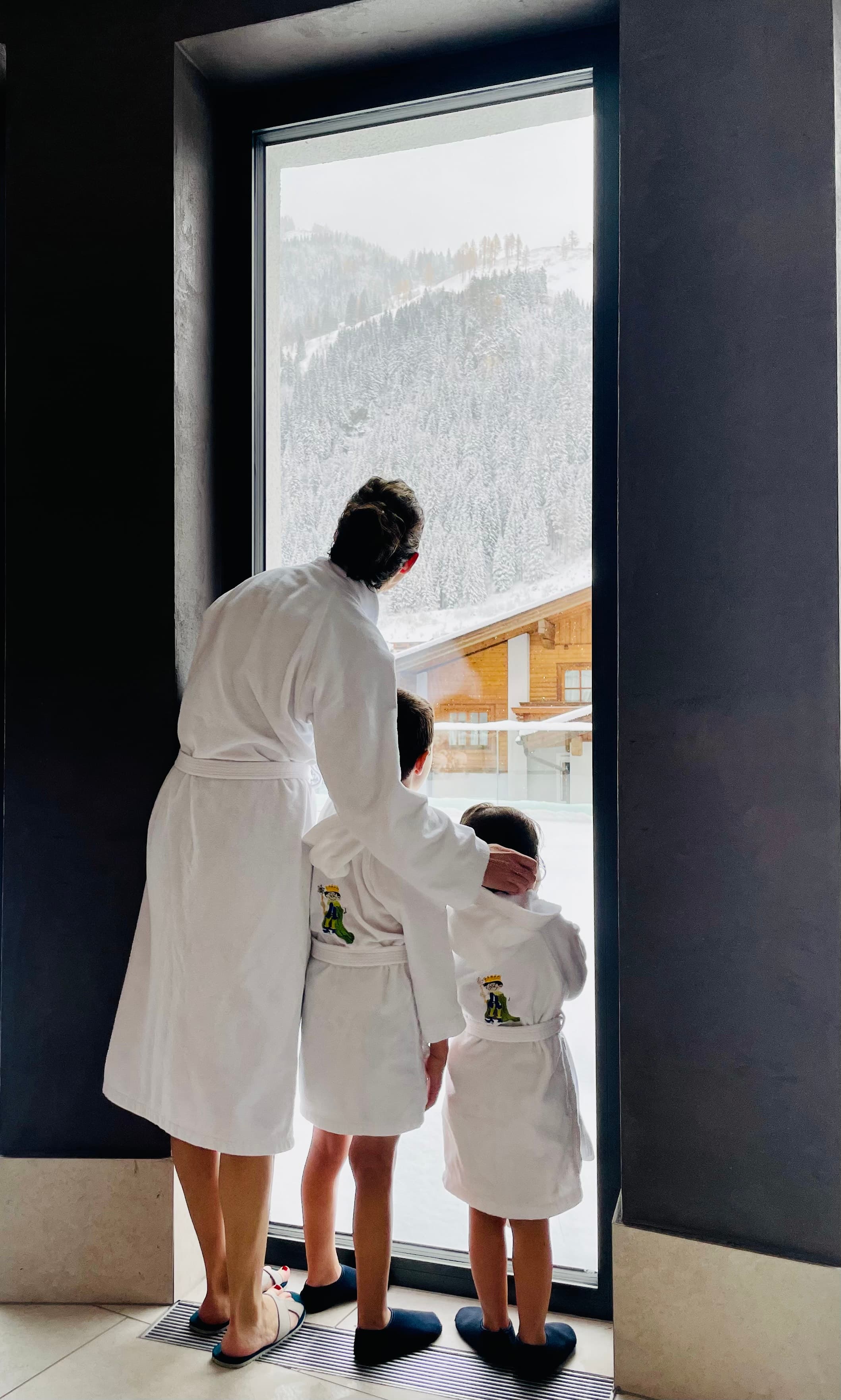 A photo of Jennifer and her two children looking out of a tall window and wearing white robes.