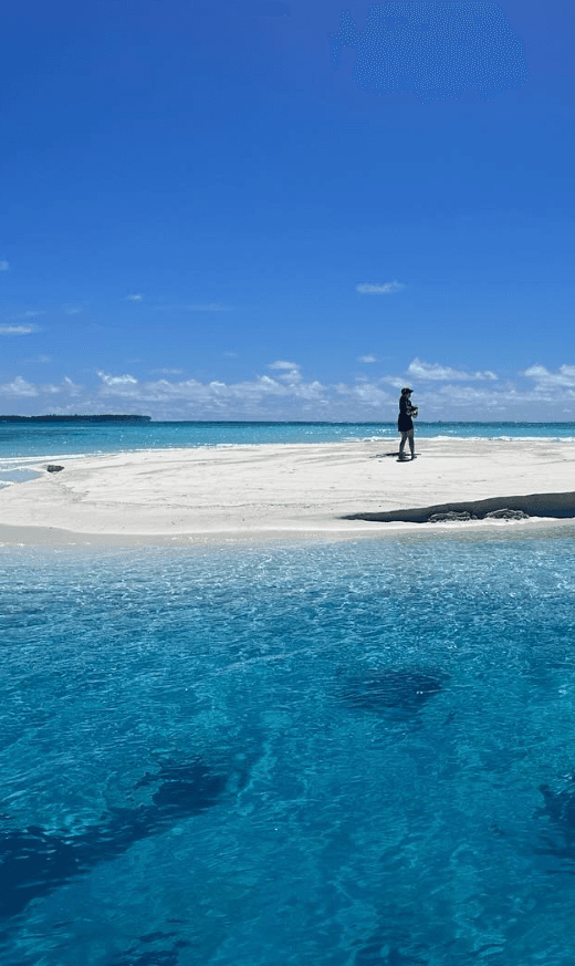 A person standing on a white sand bank surrounded by crystal clear blue water.